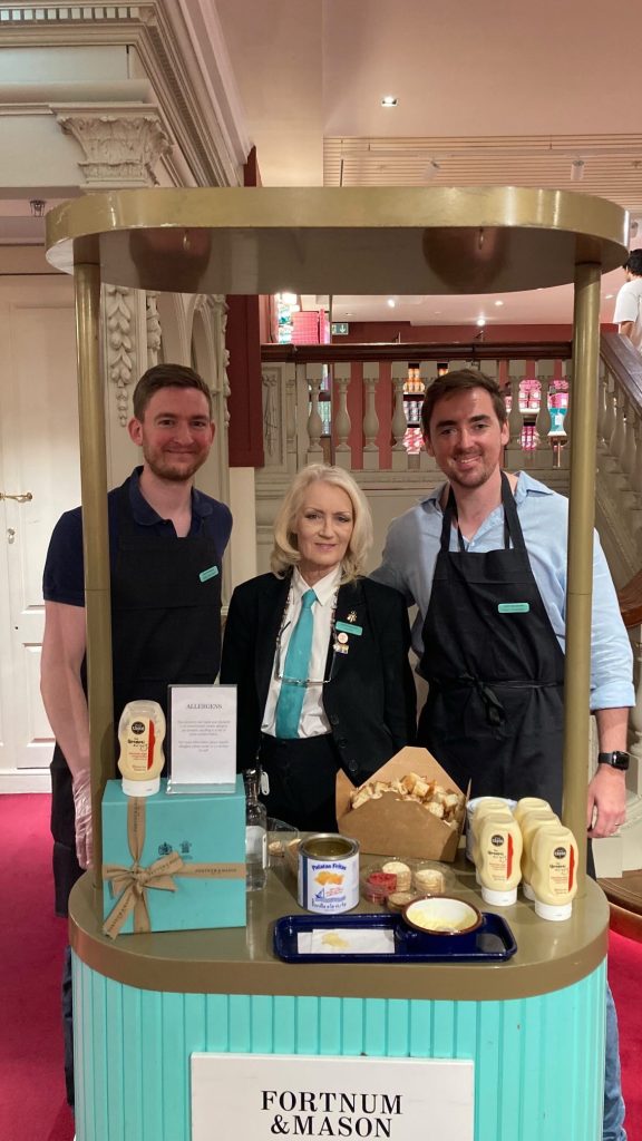 theumamibros.com Three people stand behind a Fortnum & Mason tasting stall displaying spreads, bread samples, and branded products inside an elegant shop with red carpet and a staircase in the background.