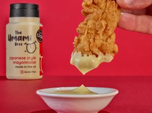 theumamibros.com A hand dips a piece of fried chicken into creamy mayonnaise. In the background, a bottle labelled “The Umami Bros Japanese style mayonnaise” highlights the rich, umami flavour on a vibrant red surface against a matching backdrop.