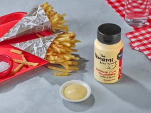 theumamibros.com Two paper cones filled with chips on a red tray, a bottle of Japanese umami mayonnaise, a small dish of creamy mayonnaise, a glass of water, salt, and a red chequered serviette on a grey surface.